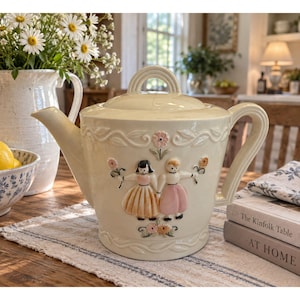 Puede incluir: Una tetera de cerámica color crema con un diseño en relieve de dos figuras sosteniendo flores, acentuado con tonos suaves en rosa y naranja. La tetera cuenta con un asa curva, un pico vertedor y una tapa con un detalle de asa arqueada.