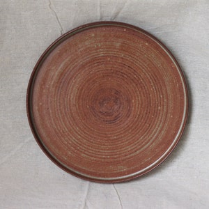 May include: A round, brown ceramic plate with a concentric circle design. The plate has a slightly raised rim and a speckled glaze.