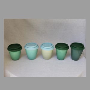 May include: Five reusable coffee cups in shades of green, blue, and cream, each with a matching lid. The cups are arranged in a row, showcasing their different colours and designs. The cups are made of ceramic.