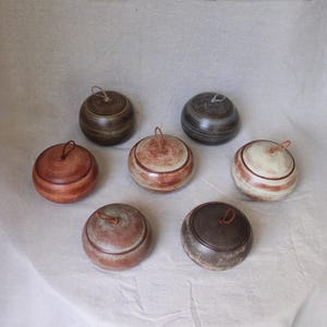 May include: Seven round ceramic lidded jars arranged in a circular pattern. Each jar features a unique glaze finish in shades of brown, cream, grey, and terracotta, with a small loop handle attached to the centre of each lid for easy opening.