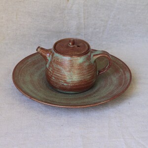 May include: A small, brown and green ceramic teapot with a lid, sitting on a matching saucer.