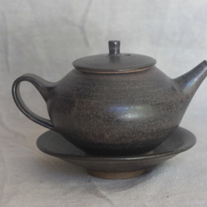 May include: A dark grey ceramic teapot with a matching saucer. The teapot has a rounded body and a spout on the side. The saucer is slightly wider than the base of the teapot.