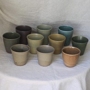 May include: A selection of ten ceramic tumblers in various earthy tones, including green, brown, and beige. The tumblers are arranged on a white surface, displaying their different hues and textures. The cups are cylindrical with a slightly tapered shape.