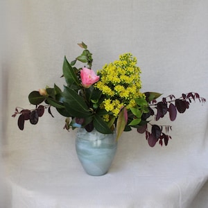 May include: A light blue ceramic vase holds a floral arrangement featuring a single pink camellia, a cluster of small bright yellow flowers, dark green glossy leaves, and several stems with deep purple-toned foliage extending outwards.
