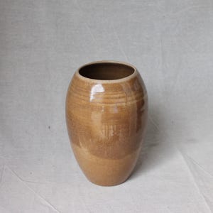 May include: A medium-sized, rounded ceramic vase with a smooth, glossy finish in shades of tan and light brown. The vessel features a wide, open circular mouth and a tapered base, showcasing subtle horizontal banding and variations in glaze colour.