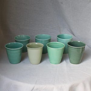 May include: Seven ceramic cups in shades of green and teal are displayed on a white surface. The cups have a simple, cylindrical shape with a slightly flared rim. The cups are handmade and have a textured surface.