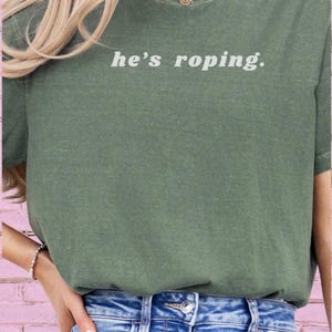May include: A green t-shirt with the text "he's roping." printed in white on the front. The shirt is being worn by a person with long blonde hair. The person is wearing blue jeans.