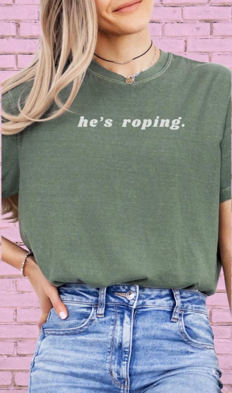 May include: A green t-shirt with the text "he's roping." printed in white on the front. The shirt is being worn by a person with long blonde hair. The person is wearing blue jeans.