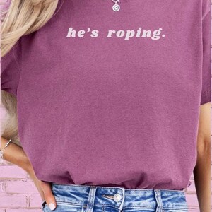 May include: A woman wearing a purple t-shirt with the text "he's roping." The shirt is a relaxed fit and has a crew neck. She is also wearing blue jeans.