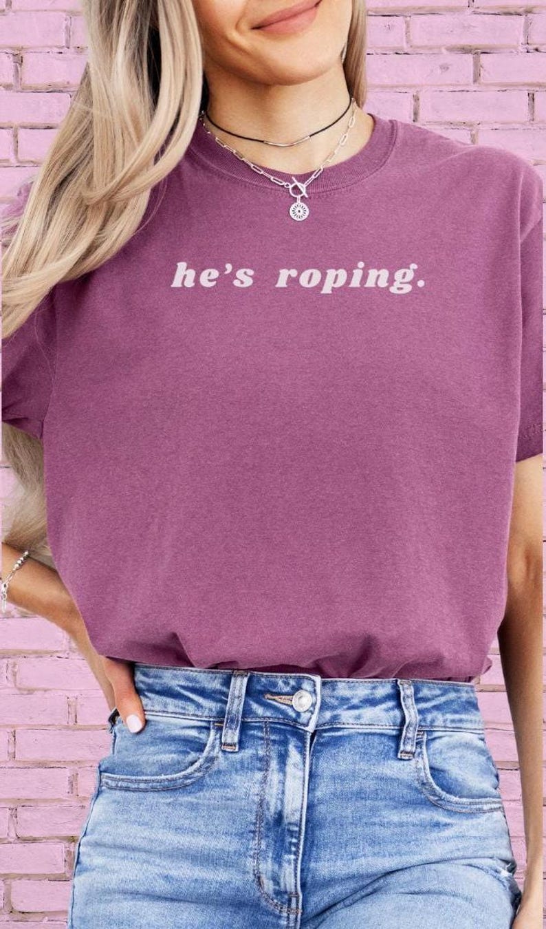 May include: A woman wearing a purple t-shirt with the text "he's roping." The shirt is a relaxed fit and has a crew neck. She is also wearing blue jeans.