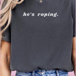 May include: A dark gray t-shirt with the text "he's roping." in white. The shirt is being worn by a person with long blonde hair and a silver necklace.