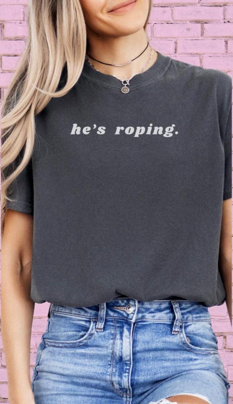May include: A dark gray t-shirt with the text "he's roping." in white. The shirt is being worn by a person with long blonde hair and a silver necklace.