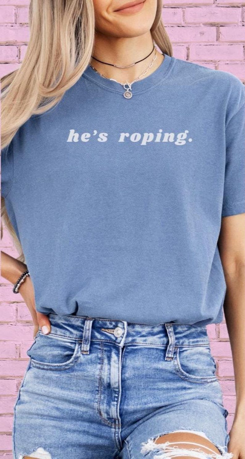 May include: A blue t-shirt with the text "he's roping." printed in white. The model is wearing the shirt with a pair of light blue jeans.