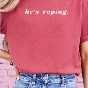 May include: A woman wearing a red t-shirt with the text "he's roping." in white. She is wearing blue denim shorts.