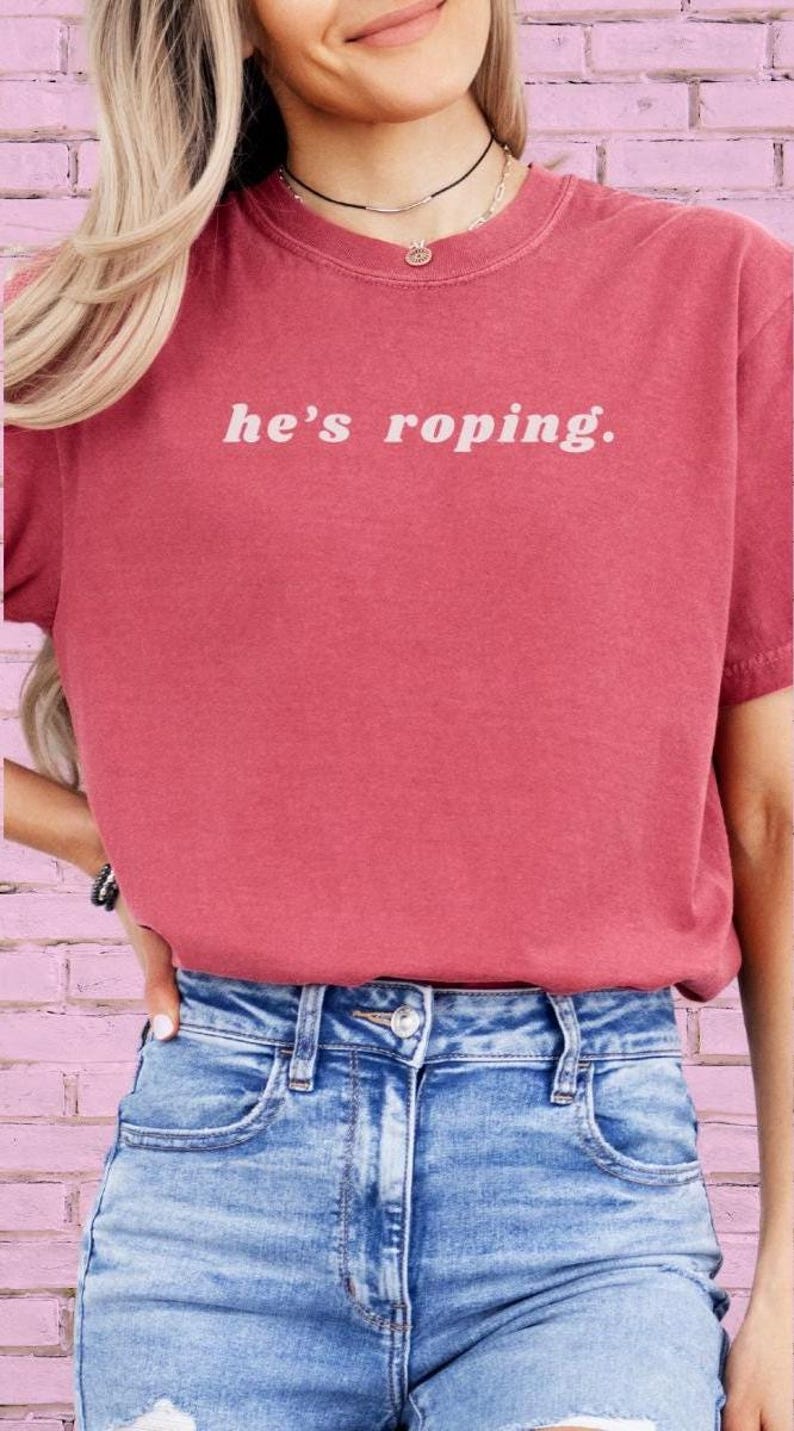 May include: A woman wearing a red t-shirt with the text "he's roping." in white. She is wearing blue denim shorts.