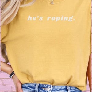 May include: A yellow t-shirt with the text "he's roping." in white. The model is wearing blue jeans.