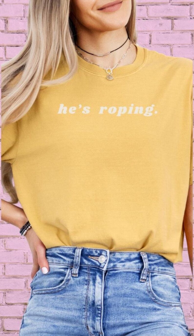 May include: A yellow t-shirt with the text "he's roping." in white. The model is wearing blue jeans.