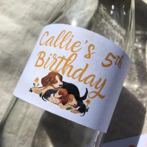 May include: A white label with the text "Callie's 5th Birthday" in gold lettering. The label features a cartoon illustration of three beagle puppies cuddling. The label is attached to a clear glass bottle.