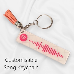 Può includere: Un portachiavi con nappa rosa con un ciondolo in acrilico trasparente che presenta un logo Spotify e un design a codice a barre rosa. Il testo "Customisable Song Keychain" è visibile sotto il ciondolo.