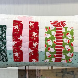 May include: Rectangular Christmas-themed pillow with a white background and quilted design. The pillow features colorful gift box patterns in red, green, and pink, with various festive prints. The pillow has a light green border.