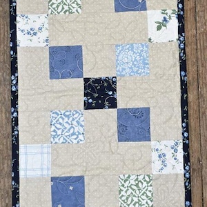 May include: A rectangular quilted table runner featuring a patchwork design. Alternating squares of beige fabric with a subtle dot pattern and various blue floral and patterned fabrics. The edges are trimmed with a dark blue floral border.