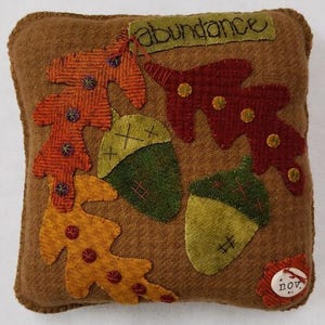 May include: A brown decorative cushion featuring appliqued autumn leaves and acorns in orange, red, and green. The word "abundance" is stitched across the top. A small tag with the word "nov" is on the bottom right.
