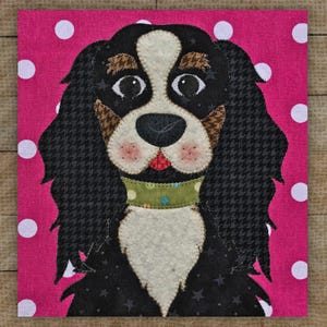 May include: A square fabric art piece featuring a Cavalier King Charles Spaniel dog. The dog has black and white fur, brown patches, and a green collar. The background is pink with white polka dots.