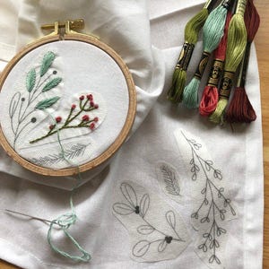 May include: A close-up of embroidery supplies on white fabric. A wooden embroidery hoop holds a partially stitched design with green leaves and red berries. Several spools of embroidery floss in various shades of green and red are visible, along with a needle and thread.