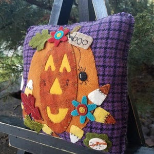 May include: A decorative Halloween pillow featuring a smiling jack-o'-lantern design. The orange pumpkin is appliqued on a purple houndstooth background. Embellishments include felt flowers, leaves, and the word "boo" on a tag.