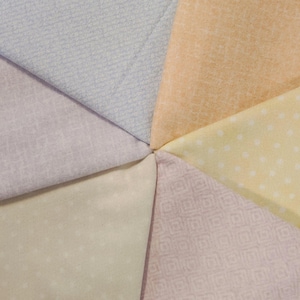 May include: A close-up of a circular arrangement of fabric swatches in pastel colors. The fabrics include light blue, peach, yellow with white polka dots, and various shades of pink and cream. The fabrics have different textures and patterns.