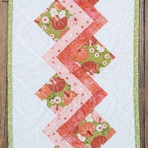 May include: A quilted table runner with a white background and a zig-zag pattern of orange, pink, and green floral fabric. The runner has a green border and measures approximately 12 inches wide.