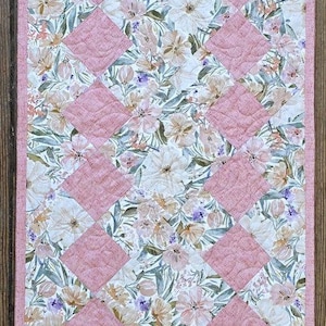 May include: A quilted table runner featuring a diamond-patterned design with alternating blocks of solid dusty pink fabric and a floral print fabric containing white, peach, and purple flowers on a cream background, finished with a pink border.