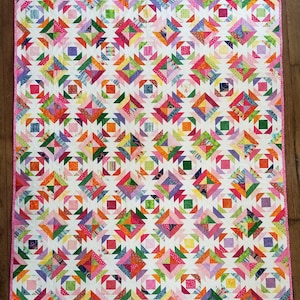 May include: A colorful quilt with a white background and a repeating pattern of geometric shapes in shades of pink, orange, green, purple, and blue. The quilt has a pink border and is likely handmade.