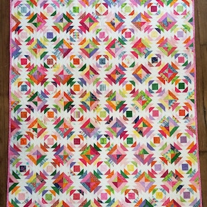 May include: A colorful quilt with a repeating geometric pattern. The quilt features squares and triangles in shades of pink, orange, green, purple, and yellow, set against a white background. The quilt is bordered with a pink fabric.