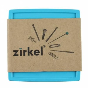 May include: A blue plastic container with a brown paper band featuring various metal fasteners. The paper band is printed with the word "zirkel" in black and green. Fasteners include a push pin, nail, screw, and paper clip.