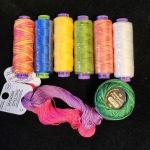 May include: A collection of colorful embroidery threads and spools. The threads include shades of pink, yellow, blue, green, and white. There is also a skein of pink and purple thread and a green spool of WonderFil Perle Cotton.