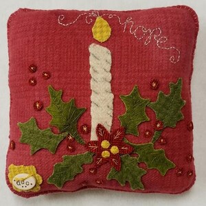 May include: A red decorative pillow with a stitched design featuring a cream-colored candle with a yellow flame, green holly leaves, and a red poinsettia. The word "hope" is stitched above the candle. A small button with the word "dec." is in the lower left corner.