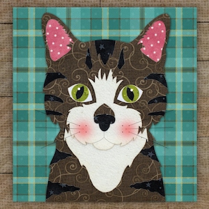 May include: A square fabric art piece featuring a stylized cat face. The cat has brown fur with a swirl pattern, green eyes, pink polka-dot ears, and a white muzzle. The background is a teal plaid pattern.