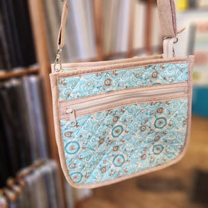 May include: A quilted crossbody bag with a light blue paisley pattern and two zippered pockets. The bag has a beige trim and strap, and silver-colored hardware. The strap is adjustable and the bag is designed for everyday use.