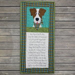 May include: A quilted wall hanging featuring a dog's portrait with brown and white fur, wearing a patterned scarf. The artwork includes a teal background and a green and patterned border. Text is embroidered below the dog.