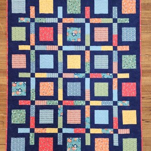 May include: A handmade quilt with a navy blue background and a grid pattern of colorful squares and rectangles. The squares feature floral and geometric patterns in shades of blue, green, yellow, and red. The quilt is bordered with a pink trim.