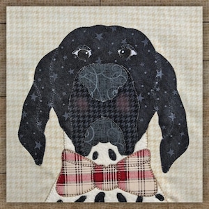 May include: A fabric square featuring a dog's face with a black and white color scheme. The dog has a black head with star accents, brown eyes, and a plaid bow tie. The background is a cream color with a small repeating pattern.