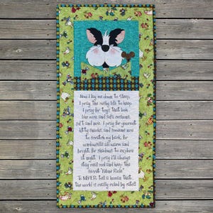 May include: A quilted wall hanging featuring a cat face applique with a green and blue floral border. The center has a poem about a cat's life, with phrases like "cushy life" and "gourmet kitty snacks."