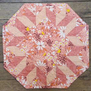 May include: An octagonal quilted placemat with a floral pattern in shades of pink, peach, and white. The design features a central starburst pattern and a border of geometric shapes. Yellow butterfly accents add a touch of whimsy.