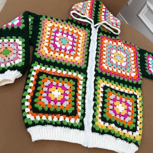 May include: A hooded zip-up jacket made of crocheted granny squares in dark green, bright orange, yellow, pink, and light blue. The garment features a white ribbed hem and cuffs, a white front zipper, and two front pockets with a matching square pattern.
