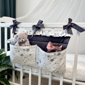 May include: A navy blue and white crib organizer with star patterns and gray bows. The organizer has two pockets and a central compartment. A teddy bear and a pink object are visible inside the pockets.