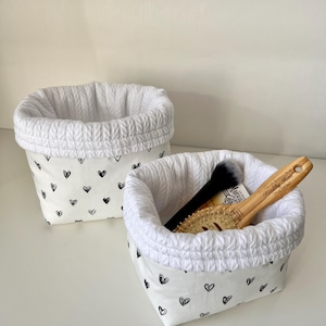 Puede incluir: Dos cestas de almacenamiento de tela blanca con un patrón de corazones negros. El borde superior de cada cesta es blanco y texturizado. Una cesta contiene un pincel de maquillaje negro y un cepillo para el pelo de madera.