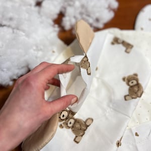 Puede incluir: Un art&iacute;culo de tela blanca parcialmente construido con un patr&oacute;n de osito de peluche, con osos y corazones marrones. Una mano sostiene el art&iacute;culo abierto, revelando el interior. Relleno blanco esponjoso visible en el fondo.