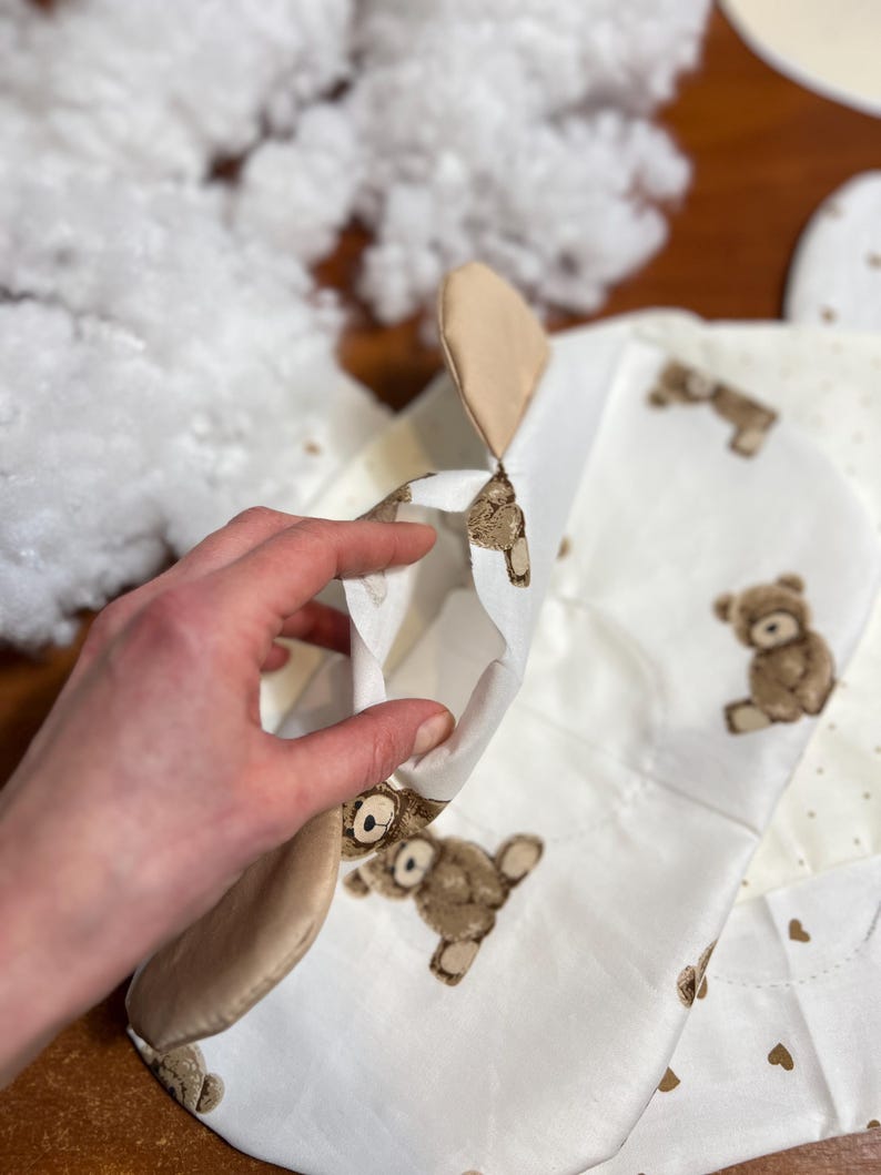 Puede incluir: Un art&iacute;culo de tela blanca parcialmente construido con un patr&oacute;n de osito de peluche, con osos y corazones marrones. Una mano sostiene el art&iacute;culo abierto, revelando el interior. Relleno blanco esponjoso visible en el fondo.