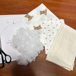 Puede incluir: Un proyecto de manualidades con trozos de tela estampados con osos y corazones, relleno blanco, una hoja de patrones y tijeras. La tela es de color crema con detalles marrones. Las tijeras son negras y plateadas.